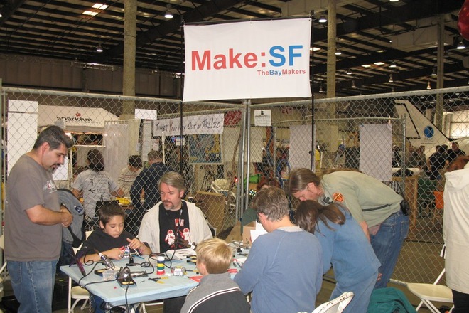 Make:SF / TheBayMakers booth at a Bay Area maker faire, with adults and children soldering and building at a shared table.