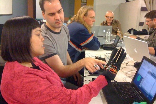 Closeup of a wired sensor glove prototype during a wearable-tech demo, with a small group gathered around open laptops.