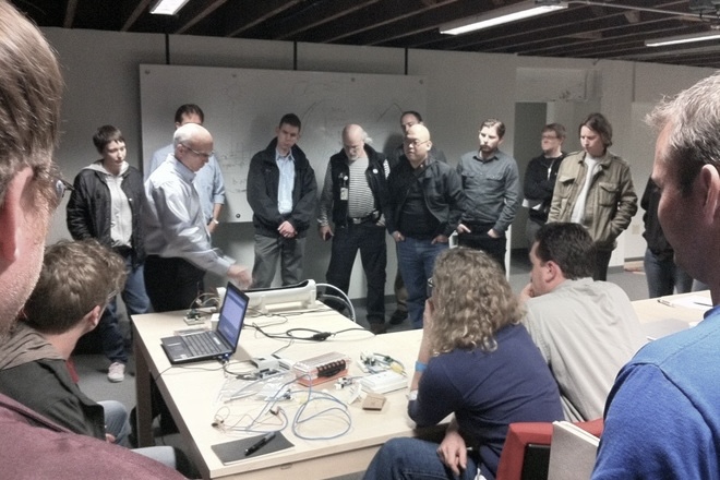 A presenter at a whiteboard walks a seated and standing group through an electronics build laid out on a workshop table.